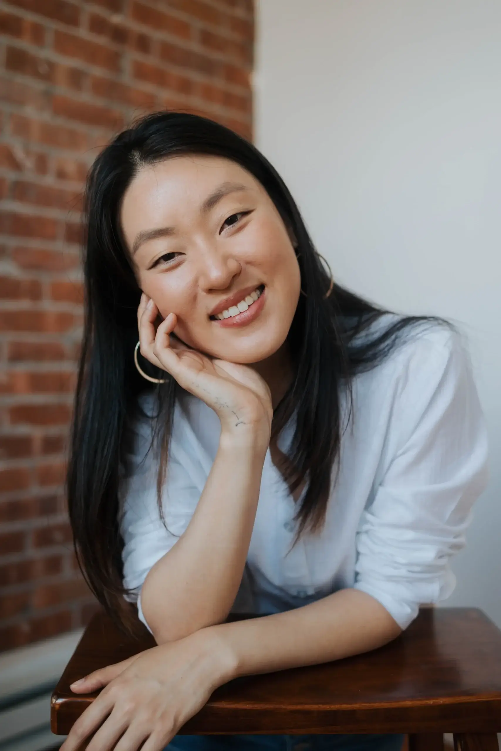 Woman with long dark hair, wearing a white shirt, smiles and rests her head on her hand in front of a brick wall at Rediscovered Psychiatry in New York.
