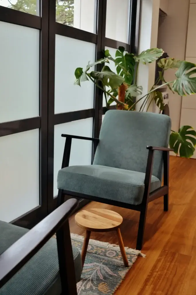 A modern lounge area in New York with green armchairs, a small round wooden table, large leafy plants, and frosted floor-to-ceiling windows on a wooden floor.