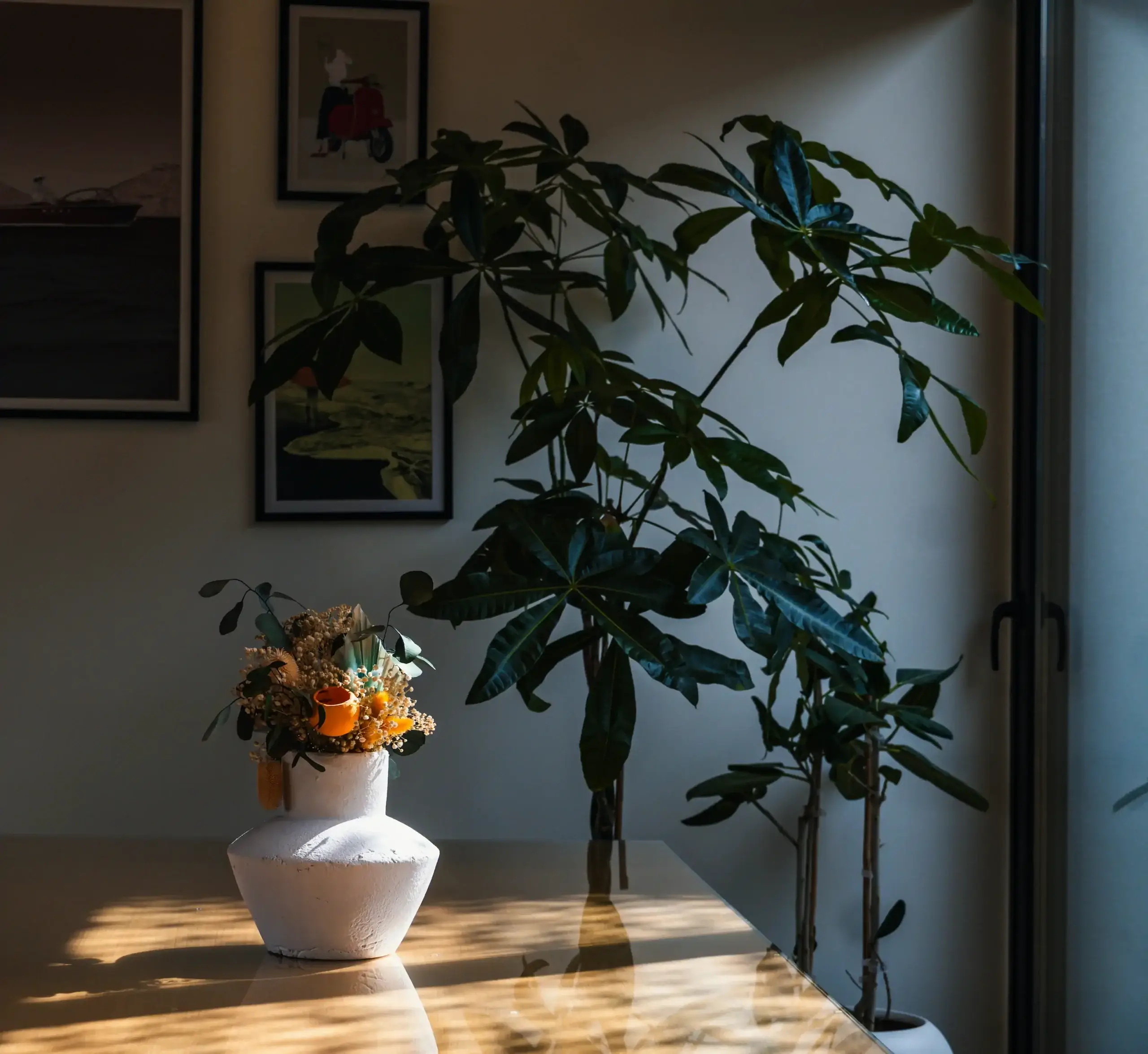 A potted plant stands by a window, while a white vase with dried flowers sits on a wooden table; framed pictures of New York hang on the wall in the background.