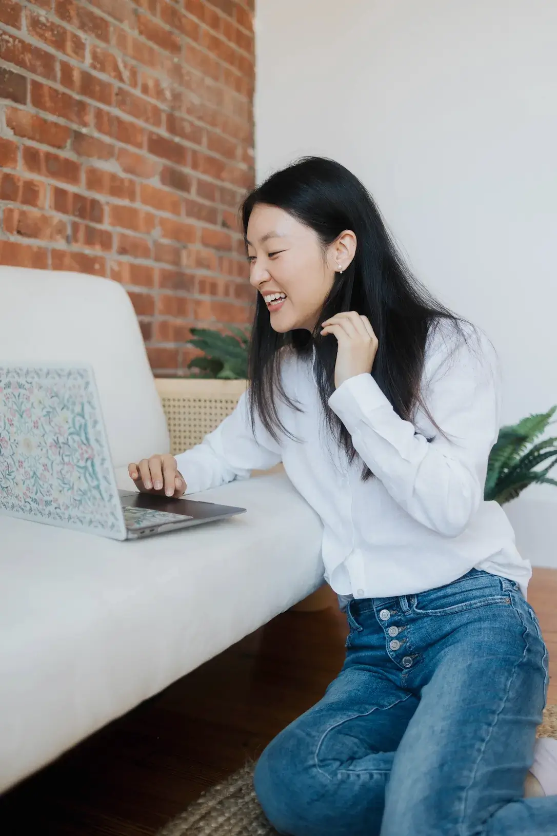 A woman smiles at her laptop beside a white sofa, enjoying a relaxed moment at home in Connecticut. Rediscovered Psychiatry inspires the decorative vibe with a plant and brick wall in view.