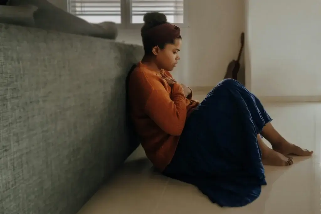 A woman sits on the floor leaning against a couch, her arms crossed and head bowed, in a softly lit Connecticut room—perhaps waiting for her Rediscovered Psychiatry appointment.