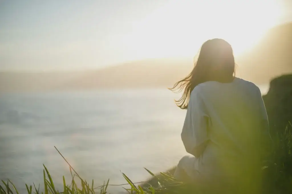 A person sits on grass near a body of water, facing the sunlight with their back to the camera, perhaps reflecting like a psychiatrist from Rediscovered Psychiatry in New York.