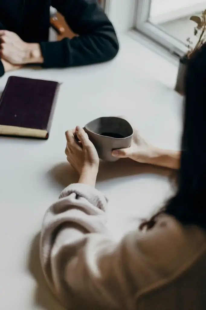 A person holding a grey mug sits at a white table with a closed dark-colored book, possibly meeting their psychiatrist in New York, and another person's hands visible in the background.