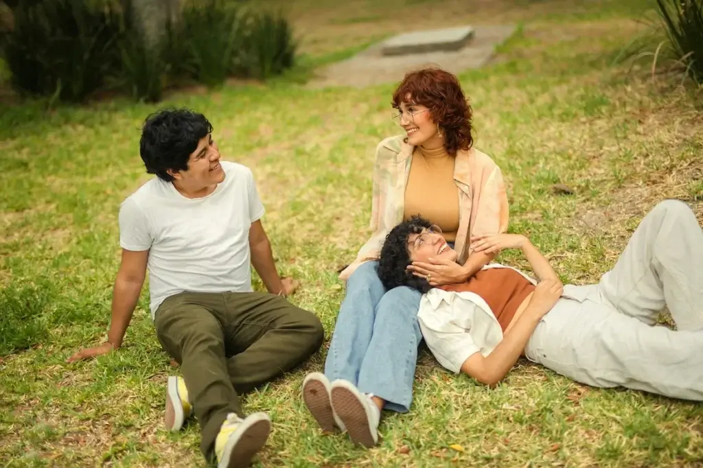 Three people relax on the grass in a New York park, smiling and interacting with each other in a cozy, casual setting.