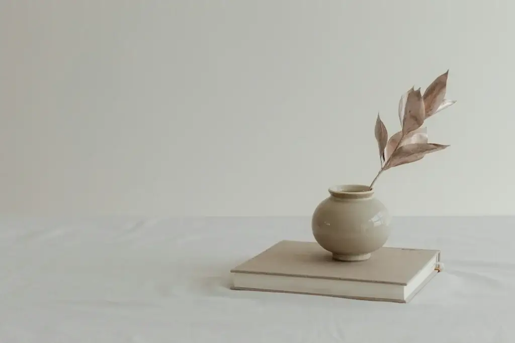 A beige ceramic vase with dried leaves sits atop two closed books on a white tablecloth, evoking the calm aesthetic of a Connecticut home against a plain light-colored background.