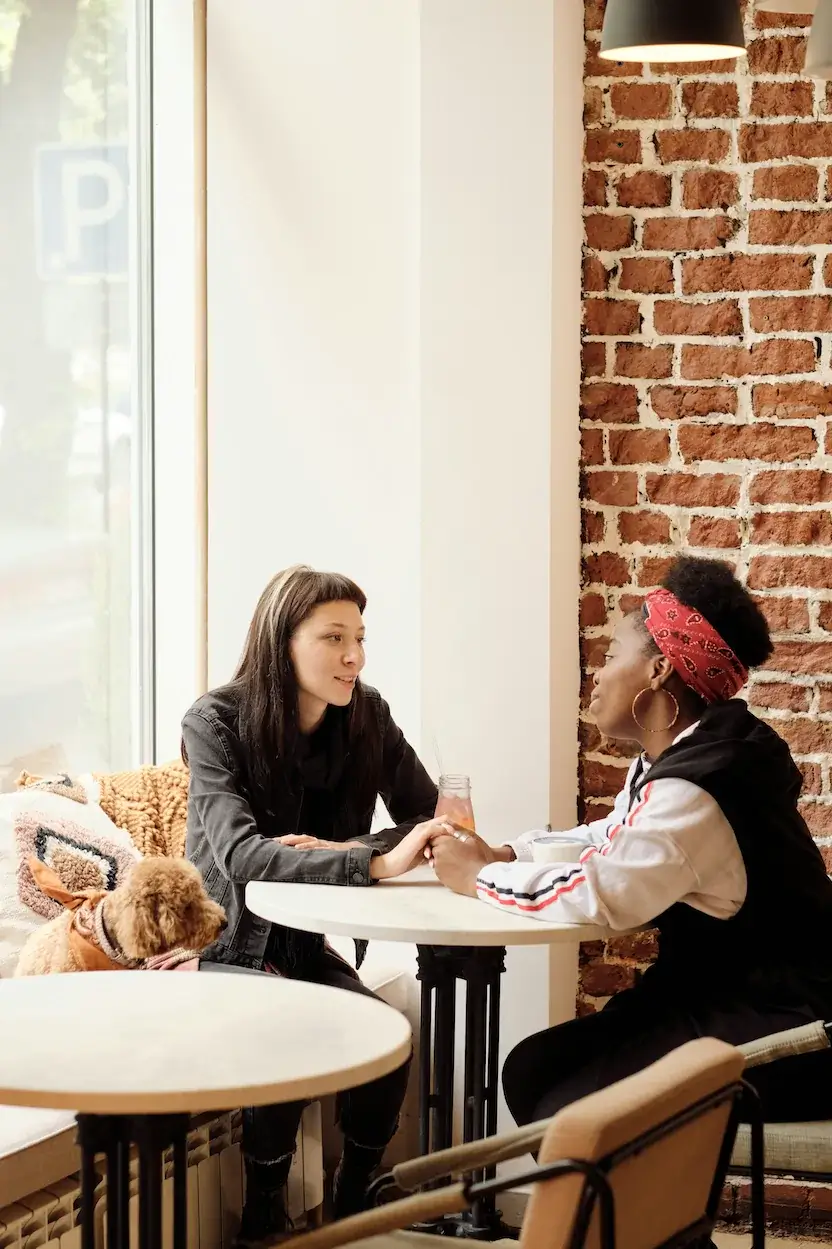 Two people sit at a round table in a cozy New York cafe, holding hands and talking. A small brown dog rests beside them near a large window with brick walls in the background.