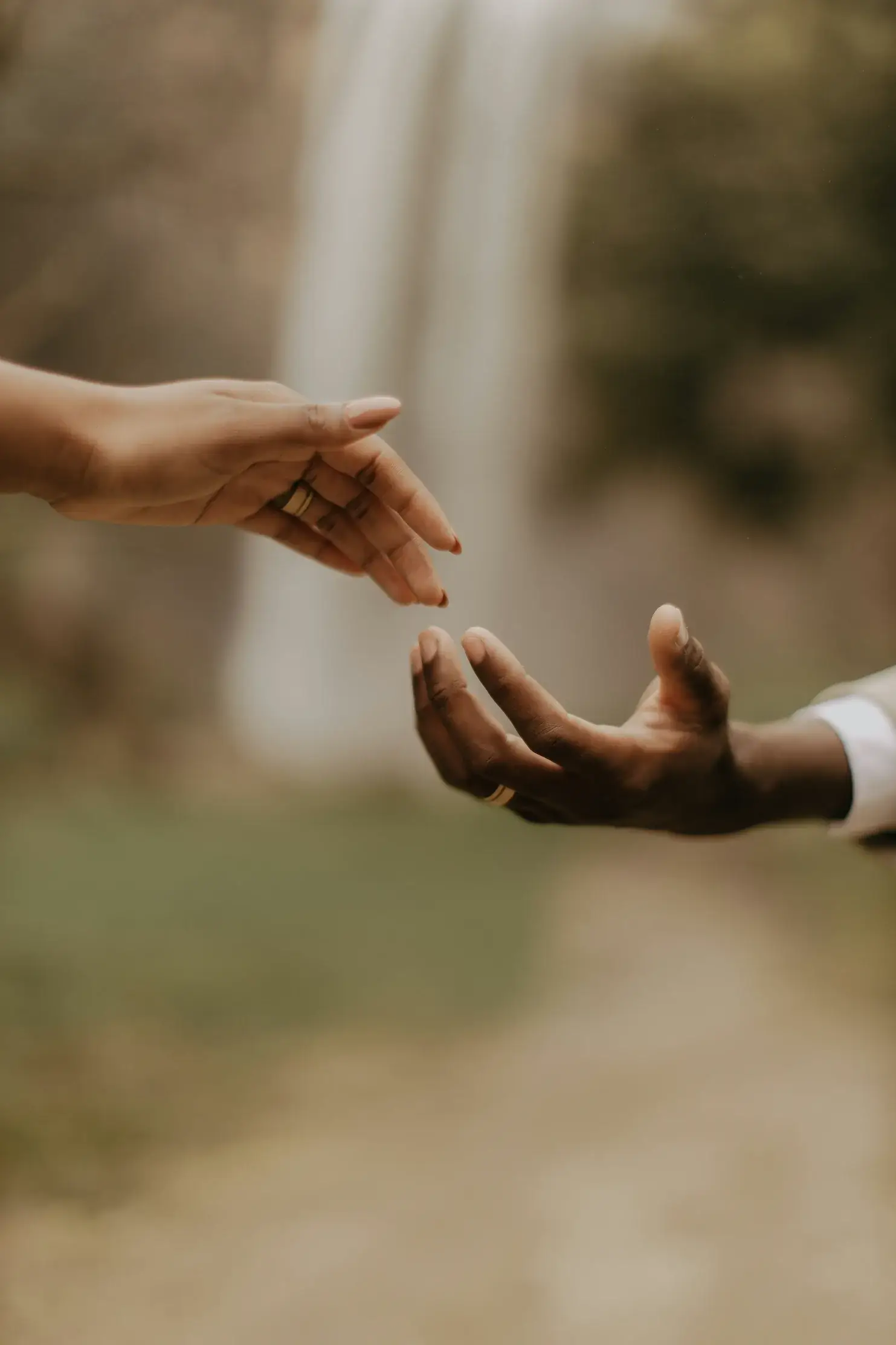 Two hands, one lighter-skinned and one darker-skinned, reach toward each other outdoors near a New York waterfall, with blurred greenery in the background.