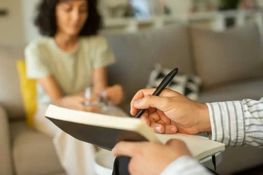 A psychiatrist writes in a notebook with a pen while another person sits on a couch in the background, appearing to talk in a Connecticut or New York office.